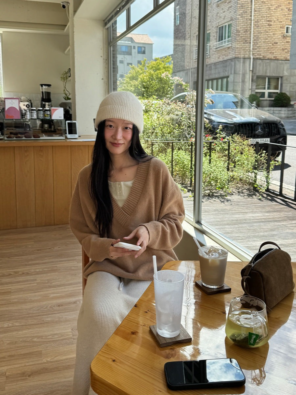 IROHI 驼色V领羊毛毛衣，模特佩戴米色针织帽，坐在咖啡馆窗边，风格简约休闲