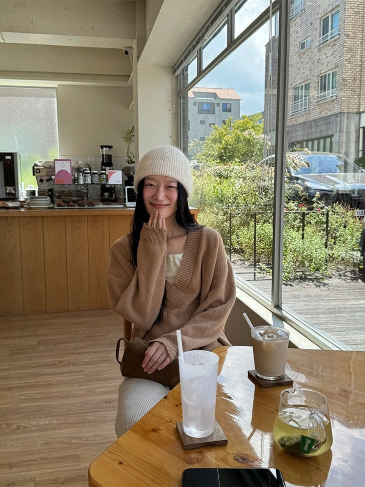 IROHI 模特身穿驼色V领羊毛毛衣，搭配白色针织帽，坐在阳光明媚的咖啡馆内。