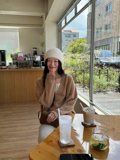 IROHI 模特身穿驼色V领羊毛毛衣，搭配白色针织帽，坐在阳光明媚的咖啡馆内。