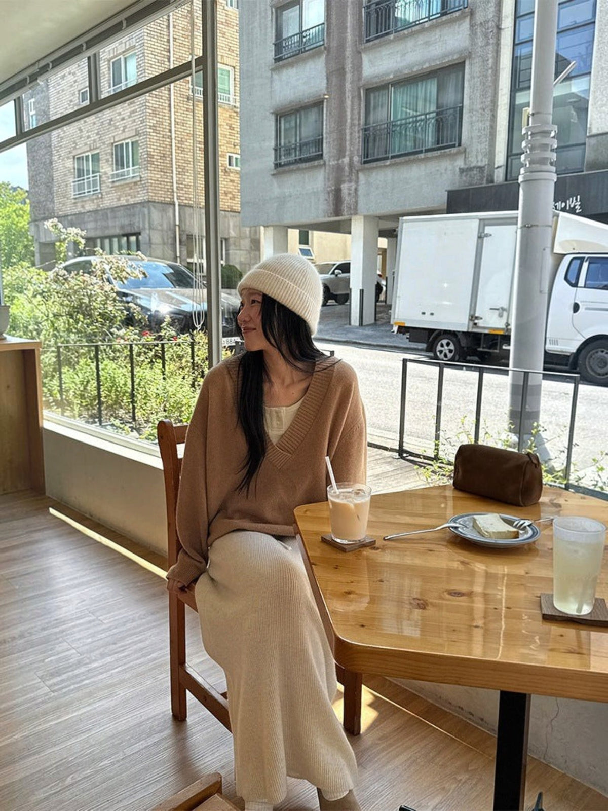 IROHI 棕色 V 领羊毛毛衣，模特身穿针织衫搭配米色长裙和针织帽，坐在咖啡馆窗边。