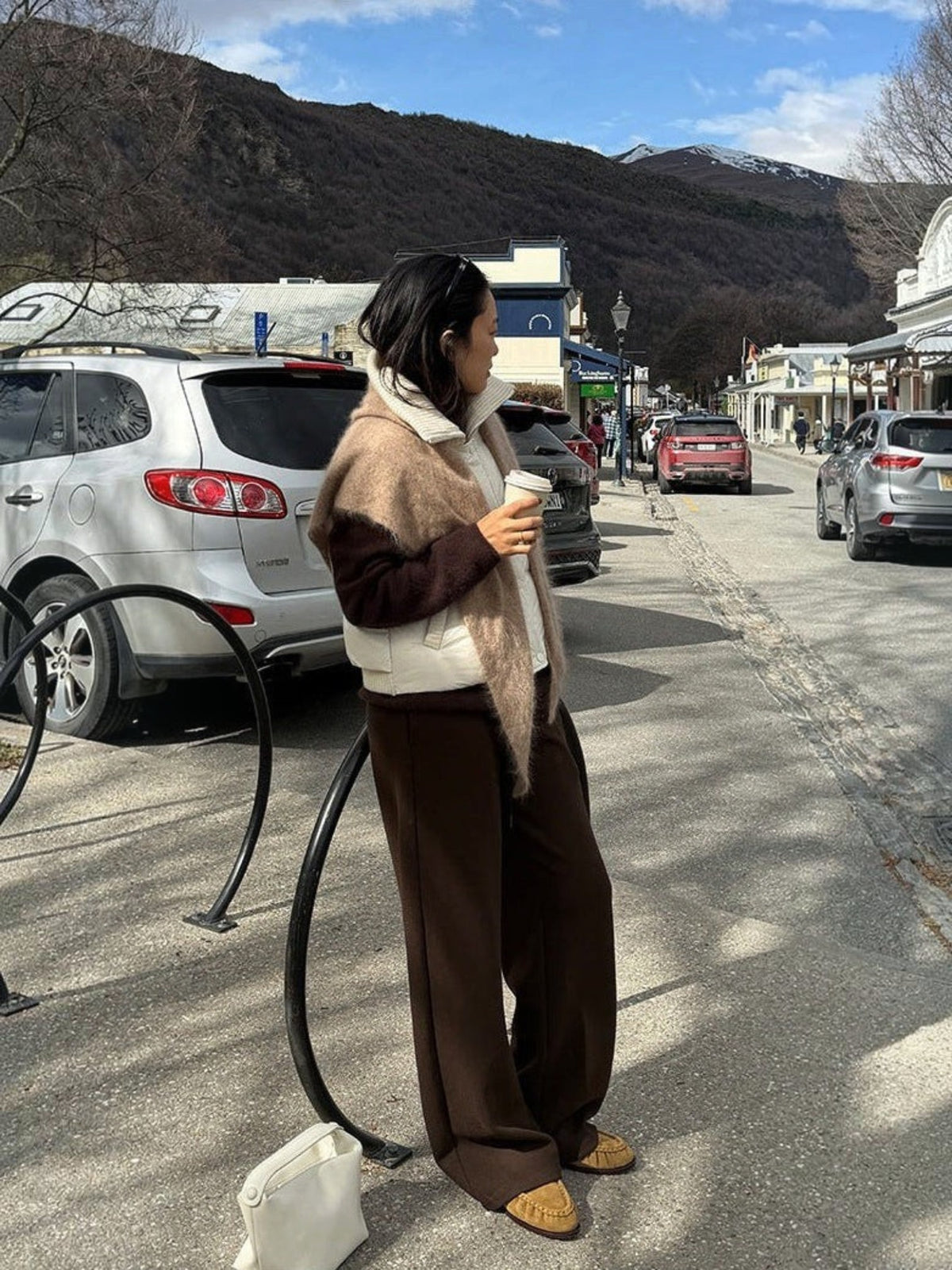 IROHI 模特身穿深棕色垂坠感阔腿裤，搭配白色羽绒背心和棕色披肩，在户外街道手持咖啡，展现简约休闲风格